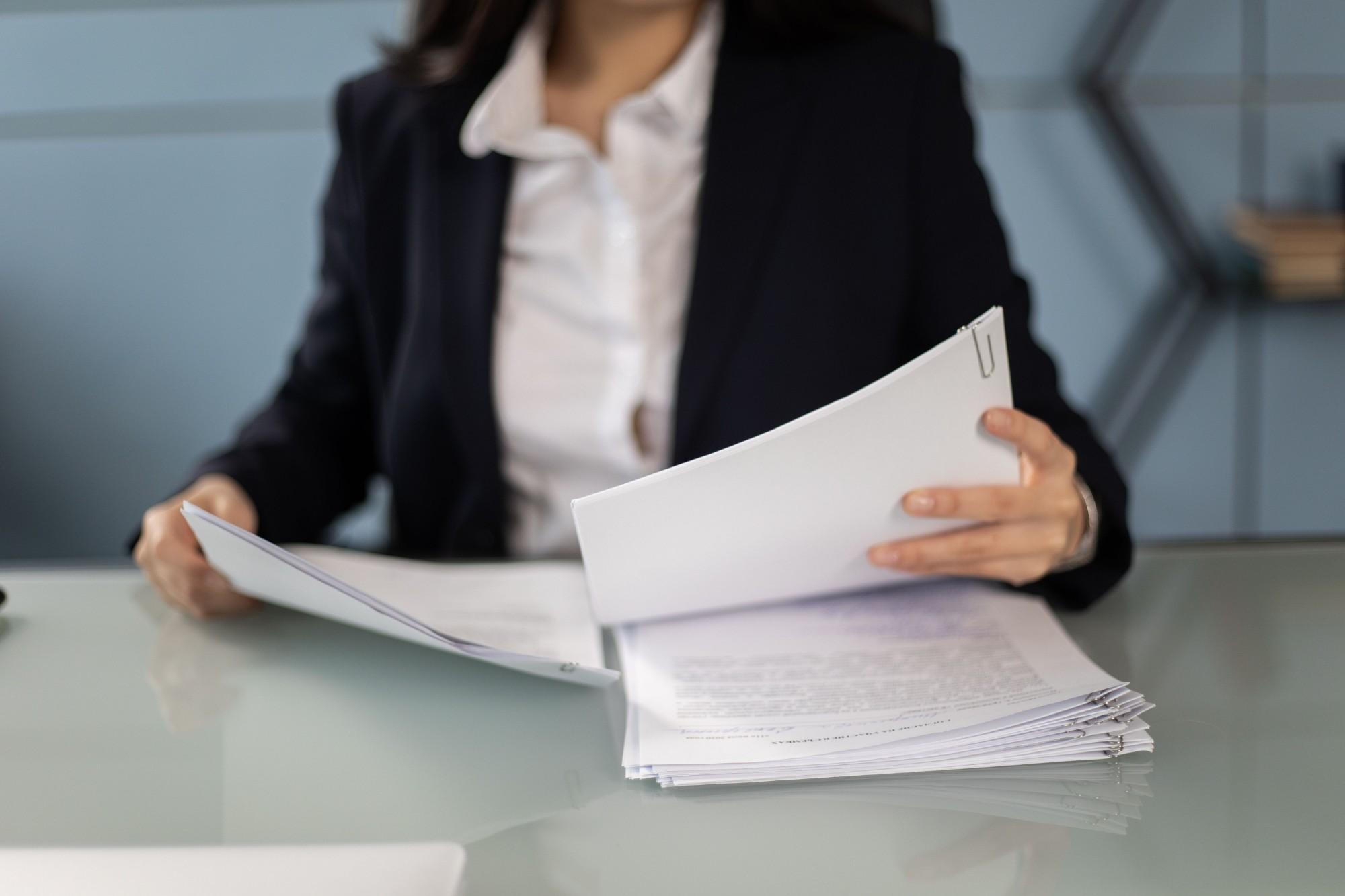 Professional in a dark blazer reviewing a stack of paper-clipped documents at a glass desk