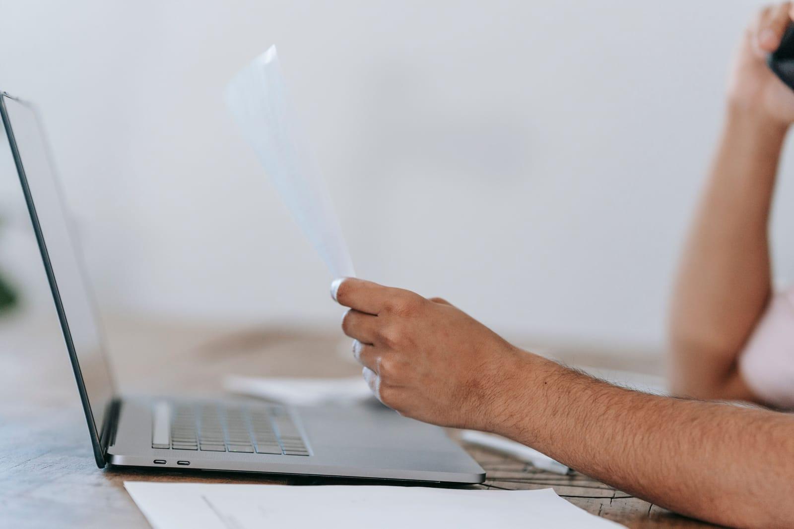 Hands holding printed documents beside an open laptop on a desk